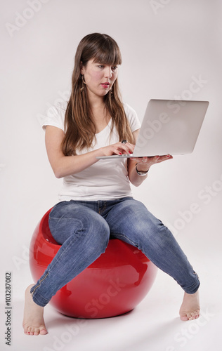 Eine junge Frau, Mädchen sitzt barfuß auf einem rotem Kunststoffsessel und hat einen Laptop in der Hand