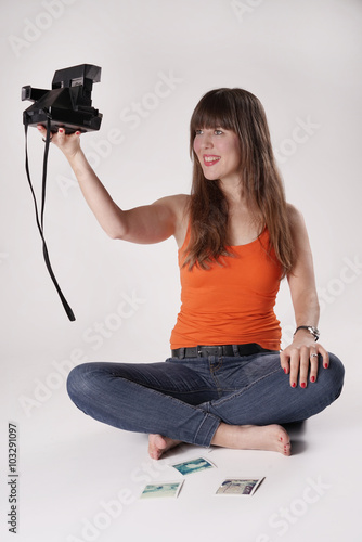 Eine junge Frau / Mädchen sitzt barfuß im Schneidersitz auf einem Boden und macht mit einer Sofortbildkamera - Polaroidkamera ein Selfie - Selbstportrait. Vor ihr liegen weitere Polaroids 
