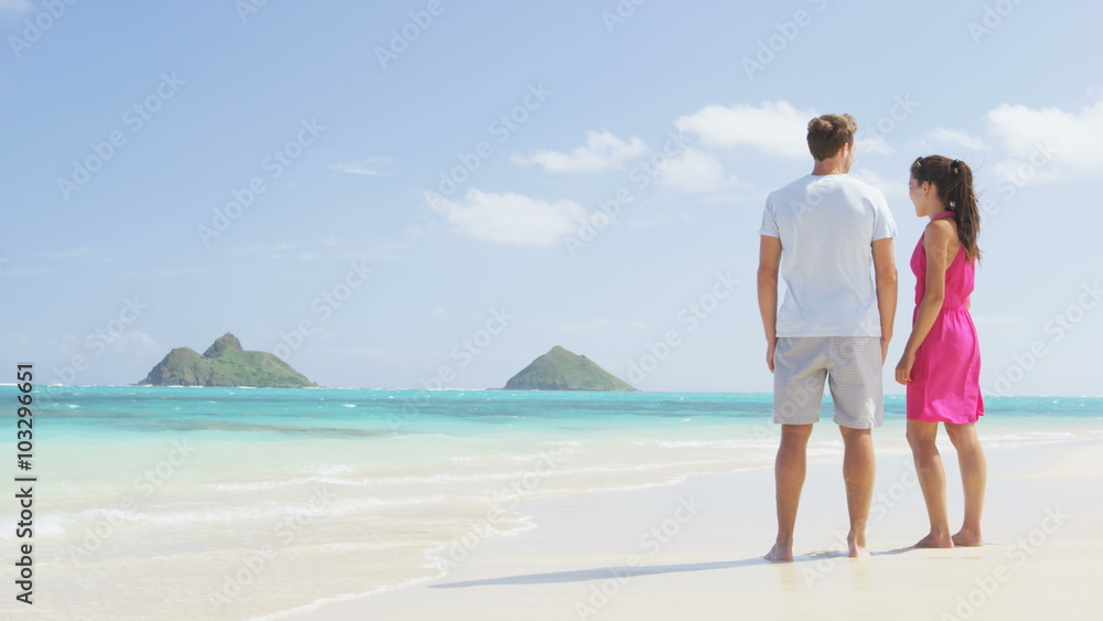 Couple on beach vacation summer holidays relaxing. Young happy people standing from behind embracing in love looking at the ocean on Lanikai beach, Oahu, Hawaii, USA with Na Mokulua Islands. Couple on beach vacation summer holidays relaxing. Young happy people standing from behind embracing in love looking at the ocean on Lanikai beach, Oahu, Hawaii, USA with Na Mokulua Islands.