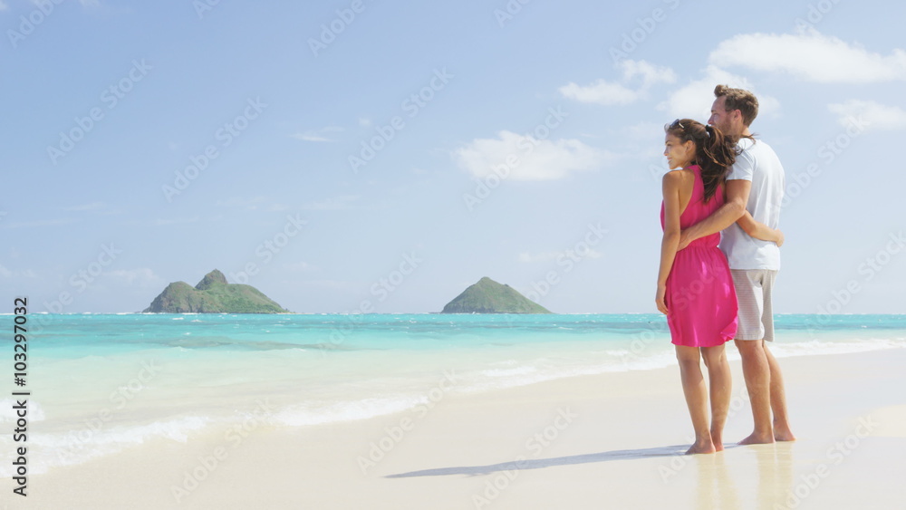 Beach summer holiday - couple on Hawaii beach vacation standing in white sand relaxing looking at ocean. Romantic young adults holding hands on Lanikai beach, Oahu, Hawaii, USA with Mokulua Islands. Beach summer holiday - couple on Hawaii beach vacation standing in white sand relaxing looking at ocean. Romantic young adults holding hands on Lanikai beach, Oahu, Hawaii, USA with Mokulua Islands.