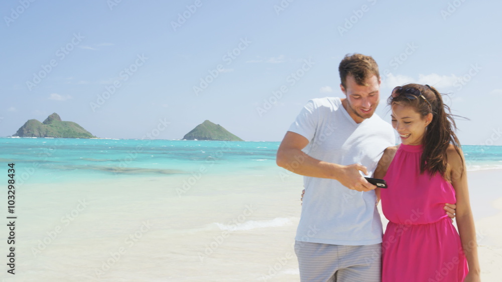 Man showing woman picture on phone app on beach using smartphone. Romantic couple in love on honeymoon having fun on outside on beach holding smart phone walking and laughing. Man showing woman picture on phone app on beach using smartphone. Romantic couple in love on honeymoon having fun on outside on beach holding smart phone walking and laughing.