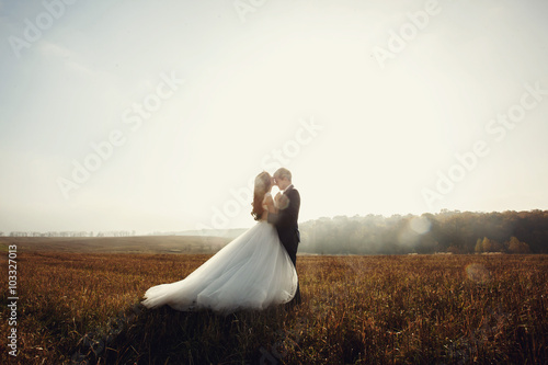 Romantic fairytale newlywed couple hug & kiss in field at sunset