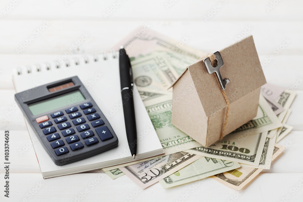 Model of cardboard house with key, calculator, notebook, pen and cash dollars. House building, loan, real estate. Cost of public utilities, insurance, rent or buying a new home concept Model of cardboard house with key, calculator, notebook, pen and cash dollars. House building, loan, real estate. Cost of public utilities, insurance, rent or buying a new home concept