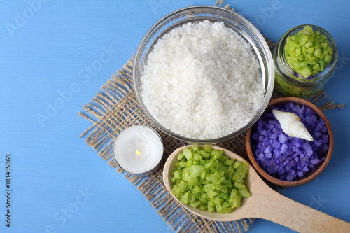 flavored green, white and purple sea salt is in a glass bowl and wooden bowl on the cloth on the blue wooden table with sea shells and a candle