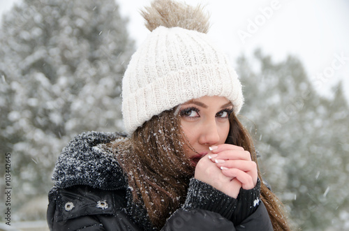 Portrait of beautiful young woman outside while its snowing, wear heavy jacket and white winter cap. Her hands are freezing and try to heat up with a breath