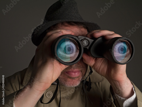 Photograph of a man in trench coat and hat looking through binoculars with huge, cartoonish eyes seen in the lenses.