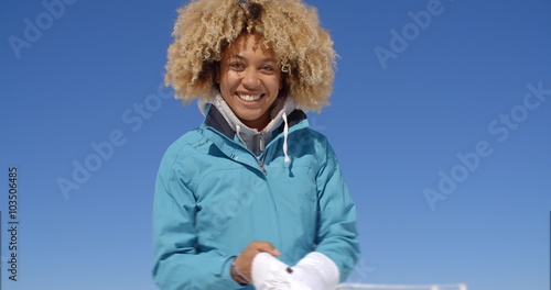 Cute young single woman in frizzy hair and winter coat with thick white ski gloves under clear blue sky