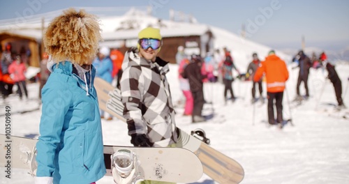 Young couple carrying their snowboards at a resort as they walk pasta ski hut and crowd of tourists on skis
