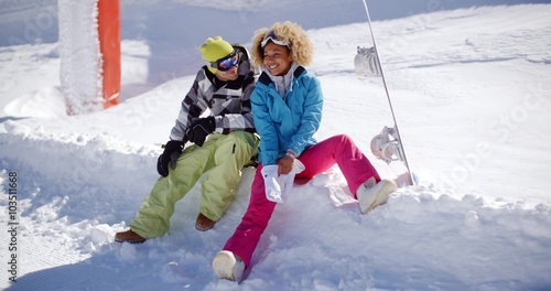 Happy young couple wearing colorful ski clothes relaxing on a snow shelf of fresh white winter snow sitting laughing and chatting in the sunshine
