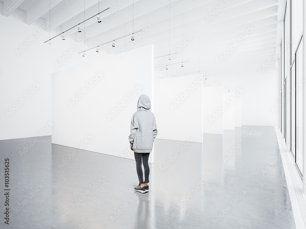 Photo of girl in empty modern gallery looking at the blank white canvas. Big windows, spotlights, concrete floor. Horizontal, mockup