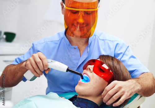 Male dentist treating female's molar with ultraviolet light in clinic. Young woman sitting in chair with open mouth wearing protective glasses. 