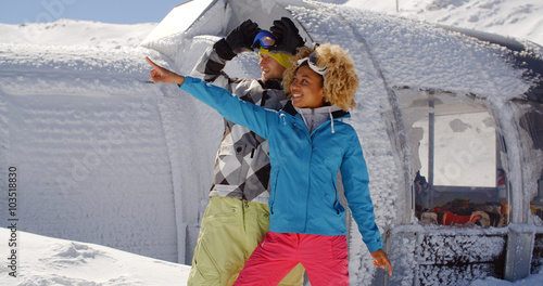 Beautiful young woman in blond curly hair pointing finger with friend at ski resort surrounded by thick white snow outside