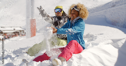 Young couple playing in the snow at a ski resort sitting in deep fresh white snow in the sunshine laughing and throwing it up into the air.