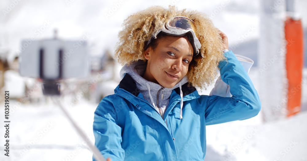 Attractive young woman with a modern afro hairdo posing in winter snow at a ski resort with her hand to her head as she smiles at the camera she holding a selfie stic with smart phone attached. Attractive young woman with a modern afro hairdo posing in winter snow at a ski resort with her hand to her head as she smiles at the camera she holding a selfie stic with smart phone attached.