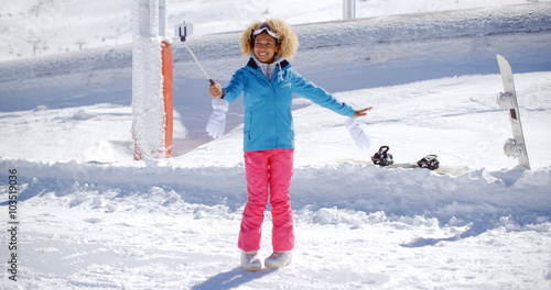 Young woman using a selfie stick as she poses with her snowboard in deep winter snow at a ski resort  with copy space