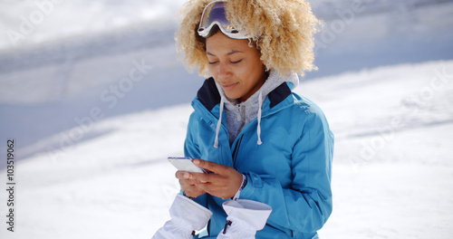 Single woman in blue snowsuit and white gloves with blond hair looking down at a phone in her hands with snowy hill in background