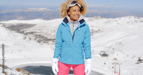 Single beautiful young woman in pink and blue ski outfit standing on mountain with big smile while looking to the side