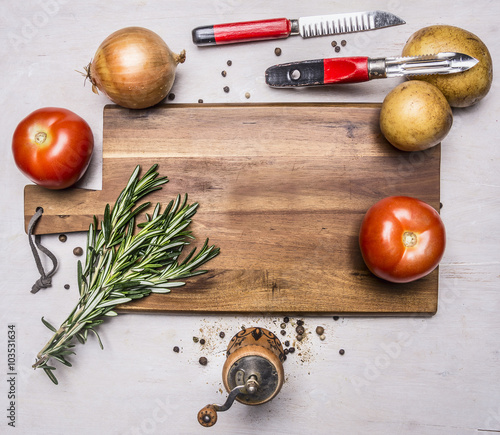 onions, tomatoes, a bunch of rosemary, potatoes, a knife for cleaning potatoes, pepper grinder posted around the cutting board on wooden rustic background top view close up photo