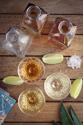 Three shots of Tequila with square bottles, lime and salt on a wooden surface viewed from above
