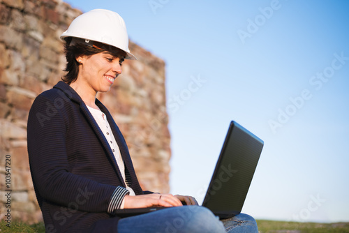Young successful woman working outdoor with laptop and safety helmet. Entrepreneur pioneer woman sitting on grass with rock wall and sky background.