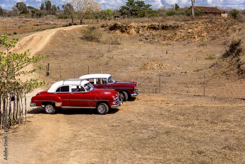 Kuba: Zwei rote US-amerikanische Oldtimer mit weißem Dach parken auf einem traditionellen Feld 