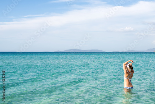Back of beautiful woman wearing blue bikini standing in the water on Mediterranean sea coast, Cesme, Ilica beach, Turkey. 