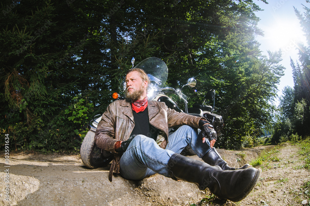 Bearded biker posing with his shiny stylish custom made cruiser motorbike. Biker is sitting on the ground near his bike wearing leather jacket, gloves and boots. Looking to the distance Bearded biker posing with his shiny stylish custom made cruiser motorbike. Biker is sitting on the ground near his bike wearing leather jacket, gloves and boots. Looking to the distance