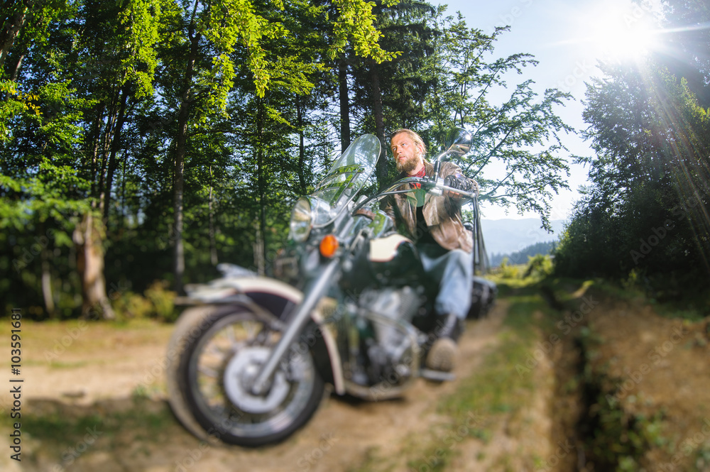 Handsome biker with beard driving his cruiser motorcycle in the forest. Man is wearing leather jacket and blue jeans. Wide angle. Tilt shift lens blur effect