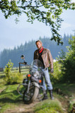 Young handsome serious man standing by his custom made cruiser motorcycle on a sunny day with forest on the background. Tilt shift lens blur effect