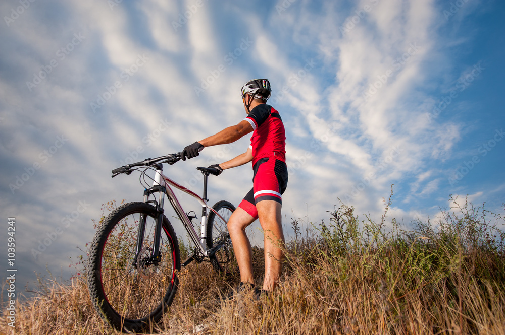 Mountain Bike cyclist in red sportswear and helmet resting with his bike against blue sky. Biker is looking into distance. Low angle view. Mountain Bike cyclist in red sportswear and helmet resting with his bike against blue sky. Biker is looking into distance. Low angle view.