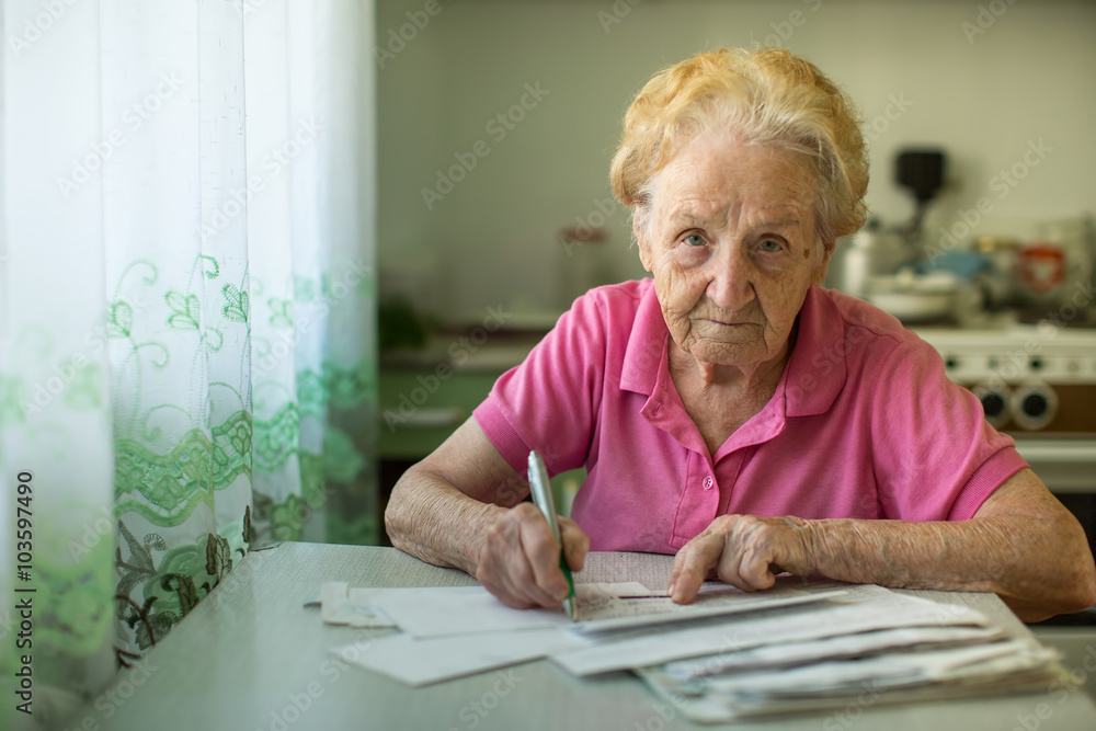 The old woman fills out utility bills sitting in the kitchen. The old woman fills out utility bills sitting in the kitchen.