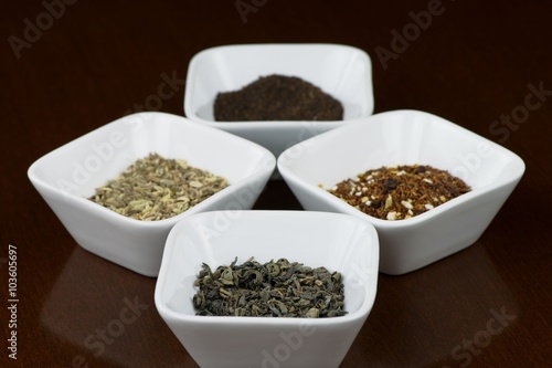 Four sorts of dry tea leaves in square plates on brown wooden table with reflection