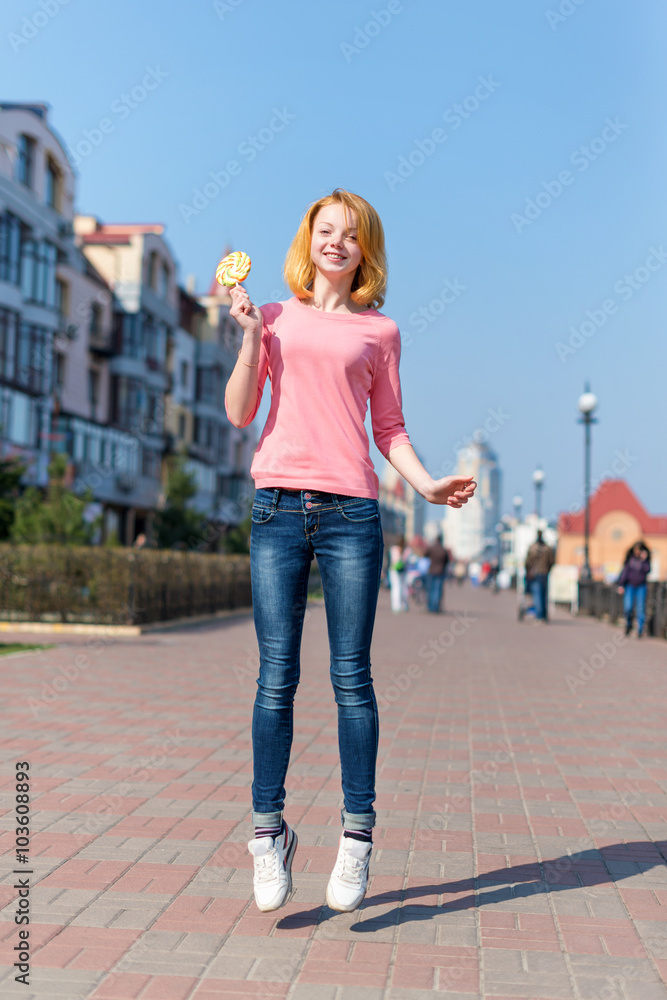 Obraz premium Redhead beautiful young woman jumping high in air over blue sky holding colorful lollipop. Pretty girl having fun outdoors.
