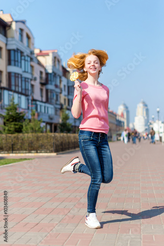 Redhead beautiful young woman jumping high in air over blue sky holding colorful lollipop. Pretty girl having fun outdoors.