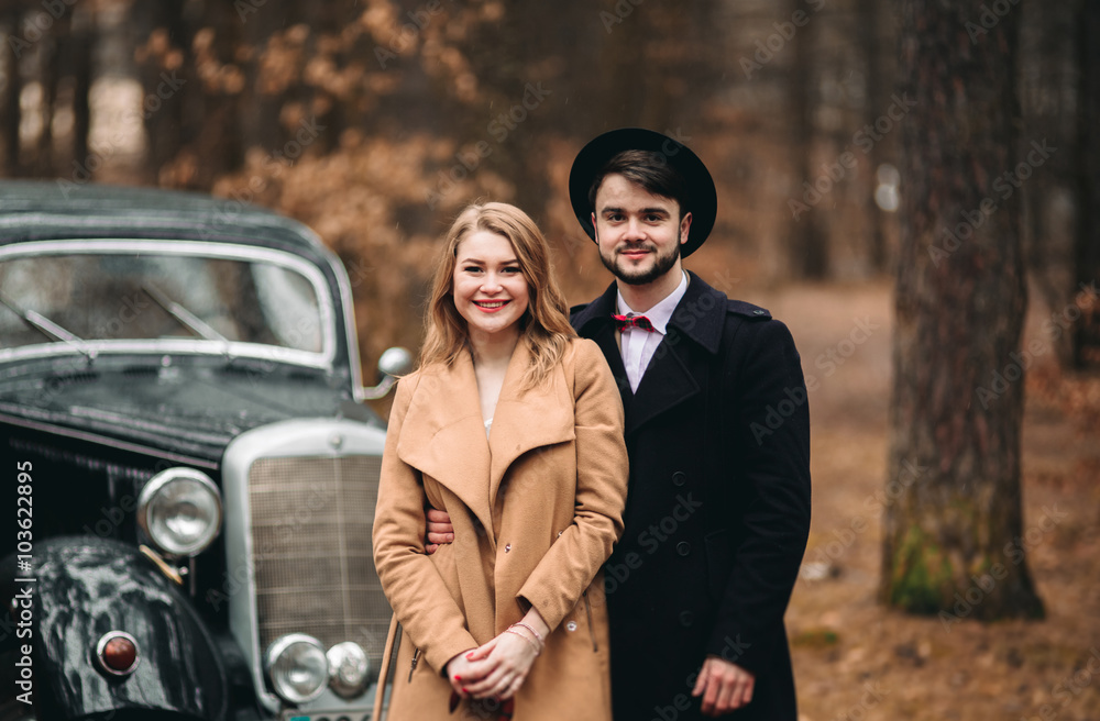 Stylish Loving wedding couple kissing and hugging in a pine forest near retro car Stylish Loving wedding couple kissing and hugging in a pine forest near retro car