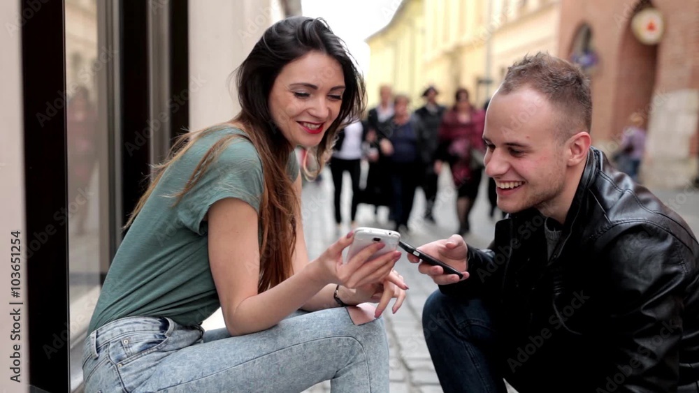 Young brown hair girl sitting on the street and talking with boy. They are looking at their smartphones and smiling. Young brown hair girl sitting on the street and talking with boy. They are looking at their smartphones and smiling.
