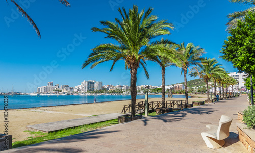 Mid morning sun, walk along the beach near the city.  Warm sunny day along the water's edge in Ibiza, St Antoni de Portmany Balearic Islands, Spain - city by the bay, row of palms lines the beach.