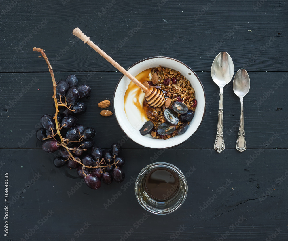 Fototapeta premium Healthy breakfast set. Bowl of oat granola with yogurt, fresh grapes, almond and honey over black wooden backdrop