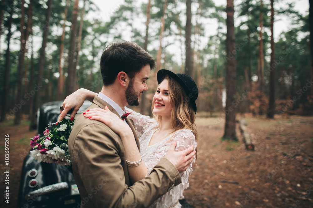 Gorgeous newlywed bride and groom posing in pine forest near retro car in their wedding day Gorgeous newlywed bride and groom posing in pine forest near retro car in their wedding day