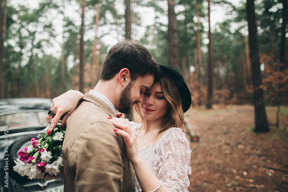 Gorgeous newlywed bride and groom posing in pine forest near retro car in their wedding day