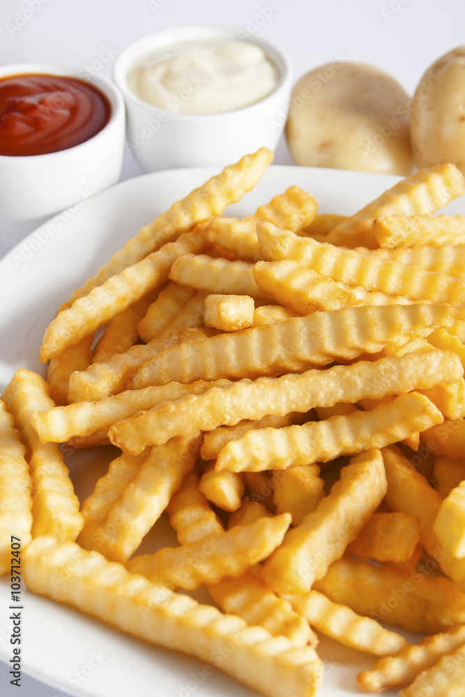 Portion of French fries (Crinkle-cut) deep fried, served on a white plate next to white bowls with mayonnaise and ketchup and fresh potato. Portion of French fries (Crinkle-cut) deep fried, served on a white plate next to white bowls with mayonnaise and ketchup and fresh potato.