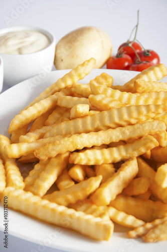 Portion of French fries (Crinkle-cut) deep fried, served on a white plate next to white bowl with mayonnaise and fresh potato.