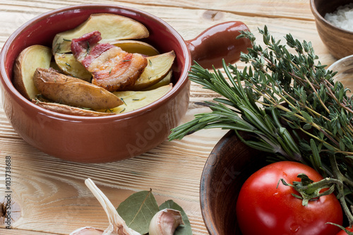 fried potatoes and bacon in ceramic forms on wooden table with clay plate with tomatoes, thyme, rosemary, sea salt, olive oil in clay jug, garlic, pepper, bay leaf