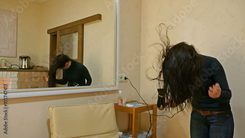 Beautiful brunette woman drying long black silky hair with hairdryer and smartens up in front of mirror in make-up dressing room. 4K UHD video footage. photo