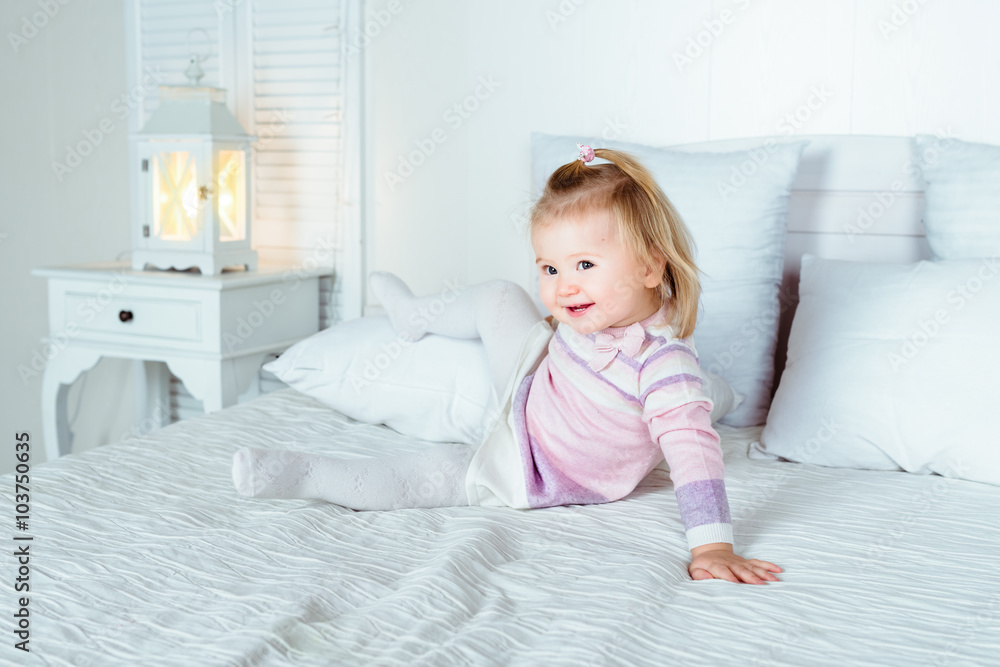 Funny and cute blond little smiling girl playing on bed in bedroom. White interior, big bed, bedside table and night lamp Funny and cute blond little smiling girl playing on bed in bedroom. White interior, big bed, bedside table and night lamp
