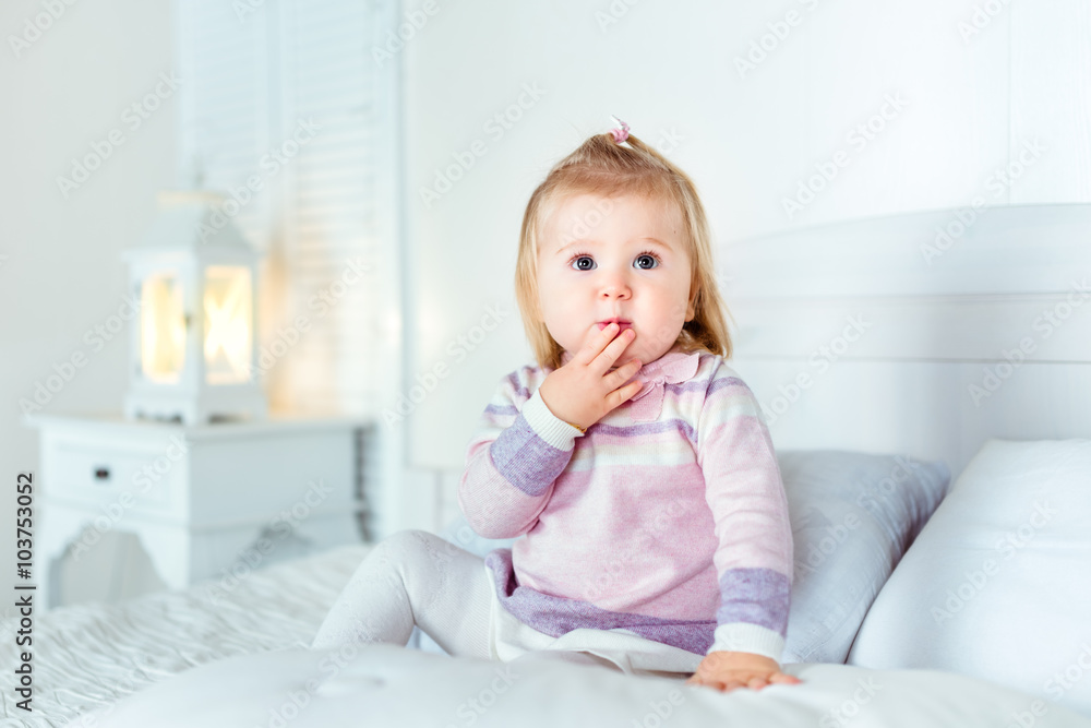 Funny amazed blond little girl sitting on bed in bedroom. White interior, big bed, bedside table and night lamp. Girl touches her mouth by hand Funny amazed blond little girl sitting on bed in bedroom. White interior, big bed, bedside table and night lamp. Girl touches her mouth by hand