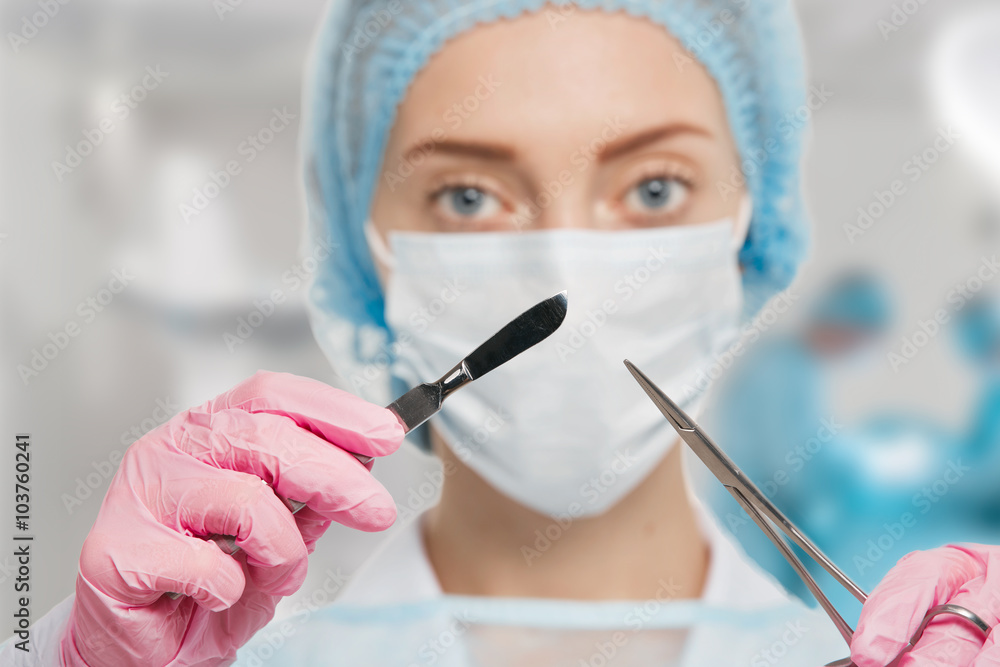 Young woman doctor with scalpel and scissors in surgery room interior Young woman doctor with scalpel and scissors in surgery room interior