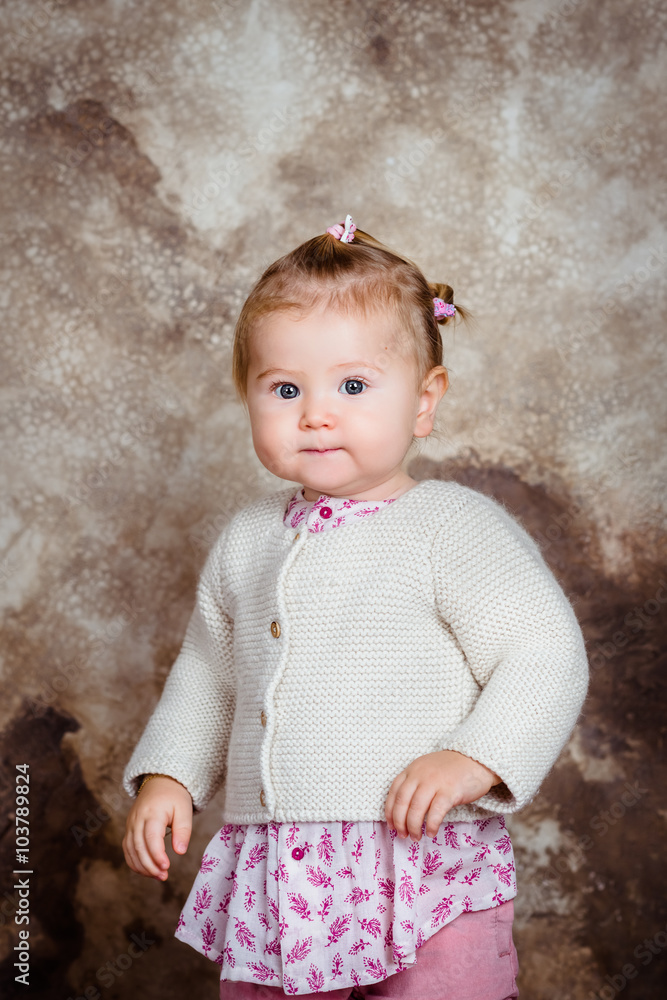 Fototapeta premium Serious little girl with blond hair and plump cheeks looking at camera. Studio portrait on brown grunge background