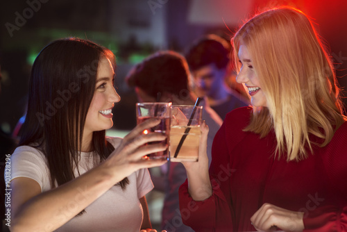 Two attractive girls clink their glasses at bar and smiling