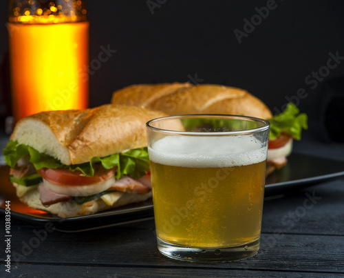 Glass of cold beer, black plate with sandwiches and beer bottle on wooden table. Fast food. Focus on beer glass.